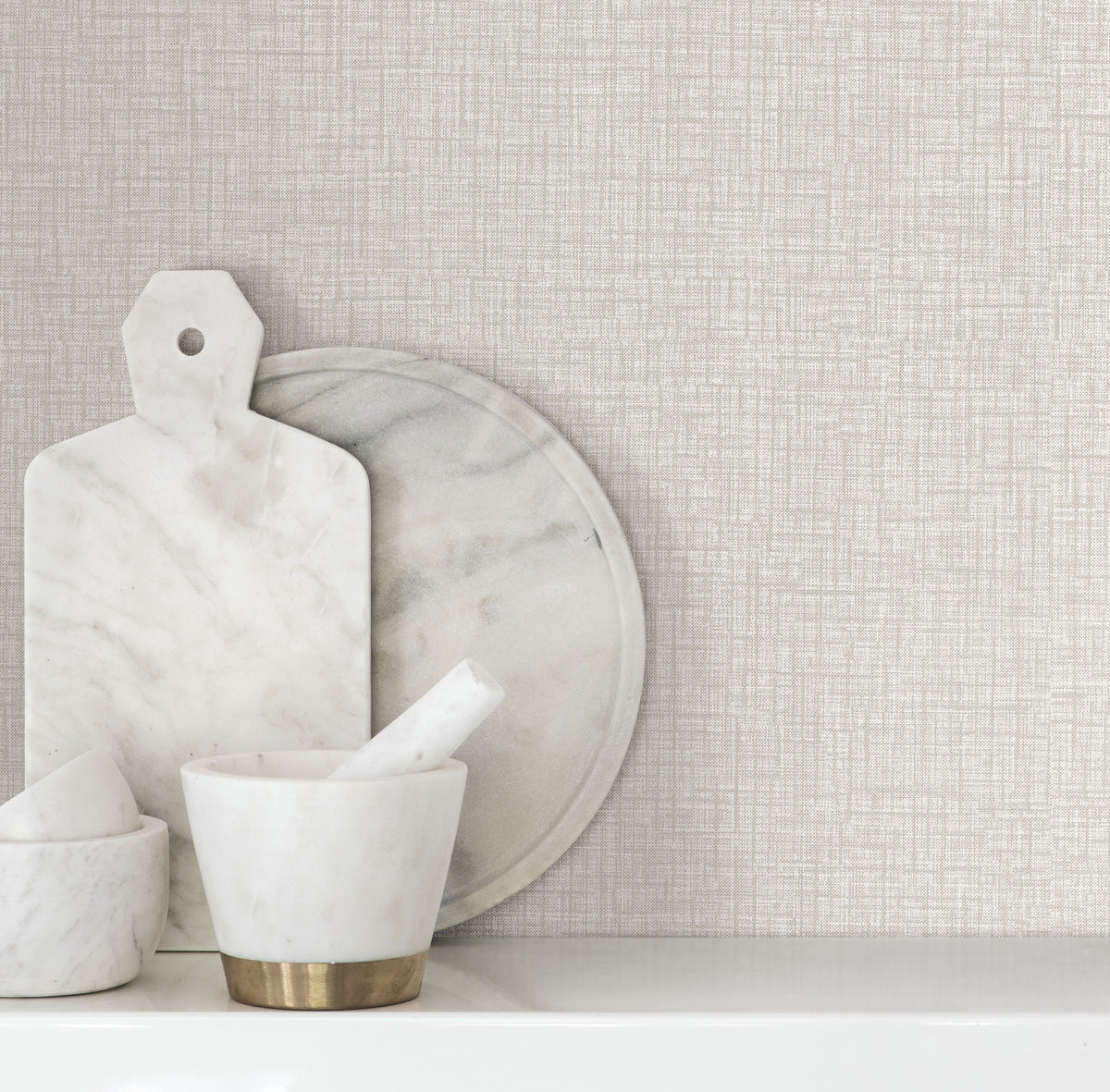 Beau Faux Fabric Wallpaper by York Wallcoverings serves as a textured beige backdrop behind a marble cutting board, round tray, and mortar and pestle arranged on a white shelf.