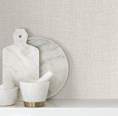 Beau Faux Fabric Wallpaper by York Wallcoverings serves as a textured beige backdrop behind a marble cutting board, round tray, and mortar and pestle arranged on a white shelf.