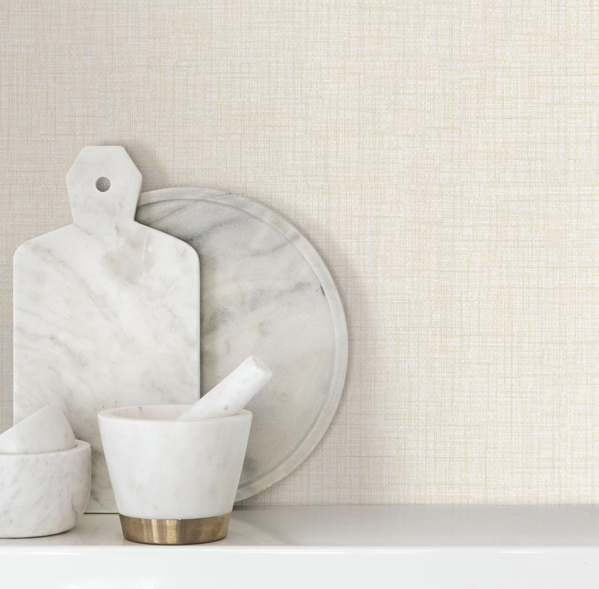 Beau Faux Fabric Wallpaper by York Wallcoverings serves as a backdrop for white marble kitchen items, including a cutting board, round tray, and mortar and pestle, arranged on a white countertop.