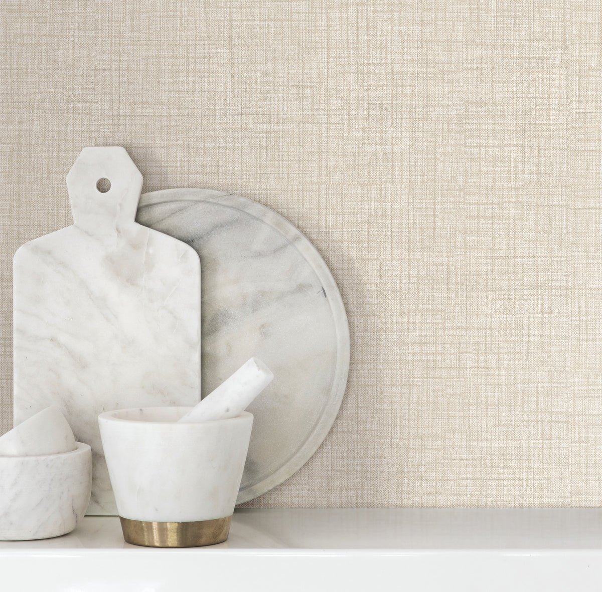 White marble kitchen items, including a cutting board, round tray, and mortar and pestle set, are arranged on a white surface against a beige Beau Faux Fabric Wallpaper backdrop by York Wallcoverings.