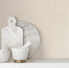 White marble kitchen items, including a cutting board, round tray, and mortar and pestle set, are arranged on a white surface against a beige Beau Faux Fabric Wallpaper backdrop by York Wallcoverings.
