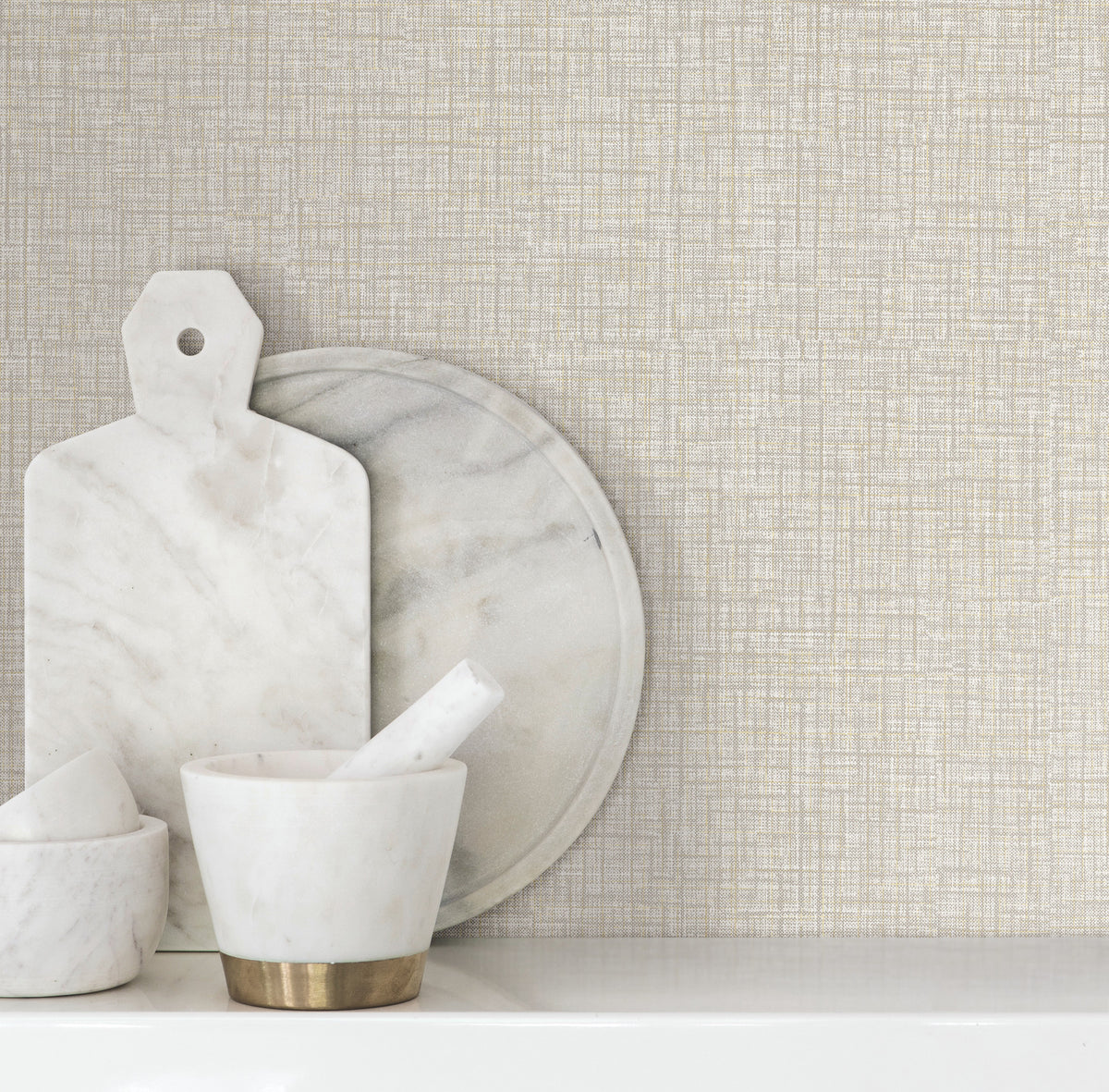 Beau Faux Fabric Wallpaper by York Wallcoverings serves as the backdrop for marble kitchen accessories, including a mortar and pestle, a round marble tray, and a marble cutting board with a handle, all arranged on a white surface.