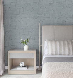 A modern bedroom featuring a light grey padded headboard, wooden nightstand, vase with flowers, and soft blue Lyle Abstract Geometric Wallpaper by York Wallcoverings. A neatly made bed is adorned with a grey blanket and striped pillow.