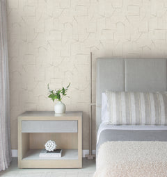 A modern bedroom featuring a gray upholstered bed, striped pillows, a beige Lyle Abstract Geometric Wallpaper by York Wallcoverings, and a wooden nightstand displaying a white vase with greenery and a geometric white decor piece on the shelf below.