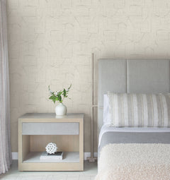 A modern bedroom featuring a light gray headboard, striped pillow, beige blanket, wooden nightstand with books, a white decorative object, and a vase with greenery is set against Lyle Abstract Geometric Wallpaper by York Wallcoverings.