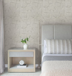 Modern bedroom featuring a light wooden nightstand, white vase with flowers, Lyle Abstract Geometric Wallpaper by York Wallcoverings, upholstered headboard, striped pillow, and a folded blanket on the bed. Soft, neutral tones create a serene atmosphere throughout the room.