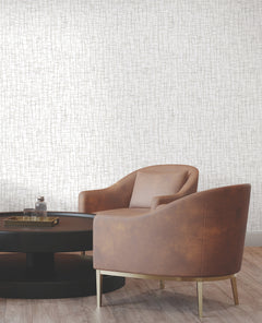 Two modern brown armchairs are placed around a round black coffee table with a glass bottle on top, set against York Wallcoverings’ Rian Linear Abstract Wallpaper and wooden flooring.