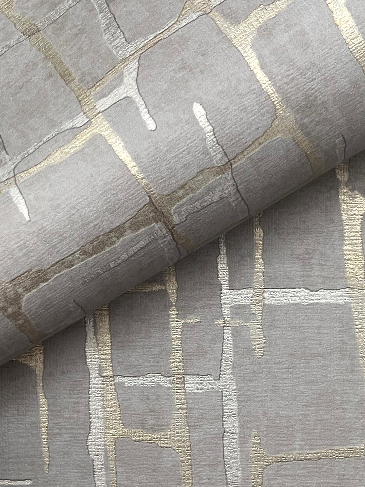 A close-up of the Rian Linear Abstract Wallpaper by York Wallcoverings showcases metallic gold and silver abstract grid patterns on textured grey paper, with one sheet partially rolled over another to highlight the wallpaper’s shimmering design.