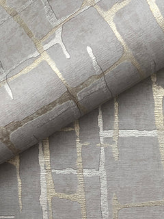 A close-up of the Rian Linear Abstract Wallpaper by York Wallcoverings showcases metallic gold and silver abstract grid patterns on textured grey paper, with one sheet partially rolled over another to highlight the wallpaper’s shimmering design.