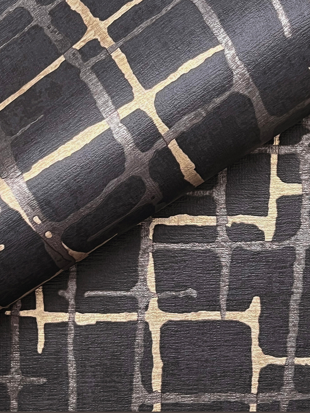 A close-up of two sheets of York Wallcoverings’ Rian Linear Abstract Wallpaper, featuring a black background with an abstract gold grid pattern and textured finish, with one sheet partially rolled over the other.