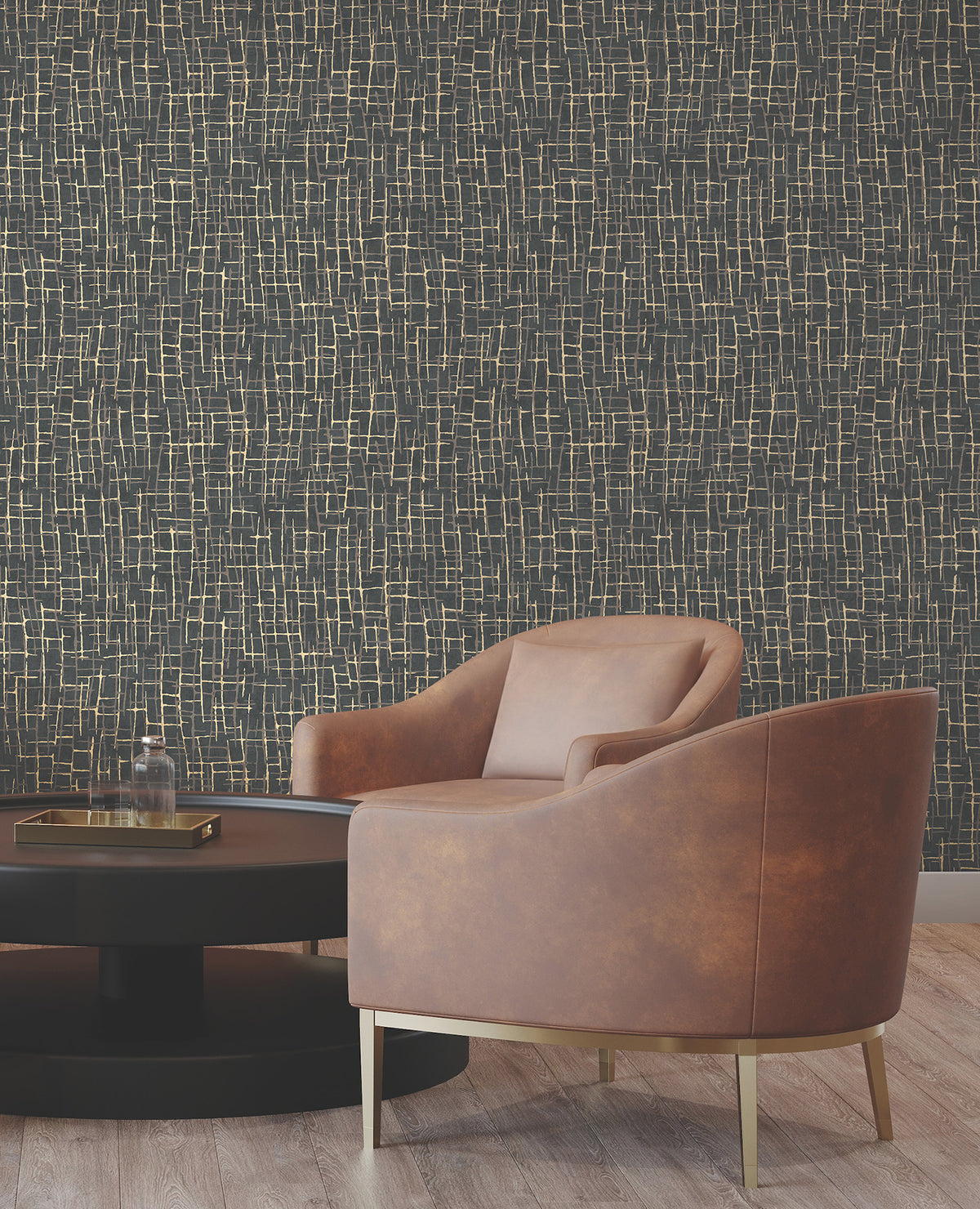 A modern brown leather chair with gold legs sits beside a round black coffee table. The background features York Wallcoverings’ Rian Linear Abstract Wallpaper in a dark hue with an abstract gold line pattern. The floor is light wood.