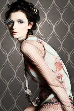 A woman in a floral top poses sideways against a wall adorned with York Wallcoverings’ Gustav Geometric Wallpaper, showcasing dramatic white feathered eyelashes and styled dark hair for a striking, avant-garde high-fashion look.