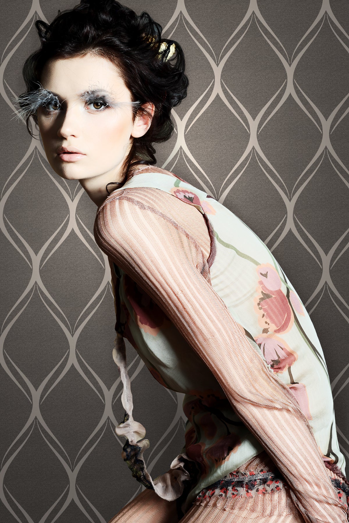 A woman with pale skin and dramatic white eye makeup poses in a sheer, floral-patterned top against a background of Gustav Geometric Wallpaper by York Wallcoverings.