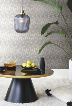 A modern living room features a round black table holding pears, a teapot, and cups. The space is decorated with the Ronald Redding Dynastic Lattice Wallpaper by York Wallcoverings, a pendant light, a large plant, and a textured rug.