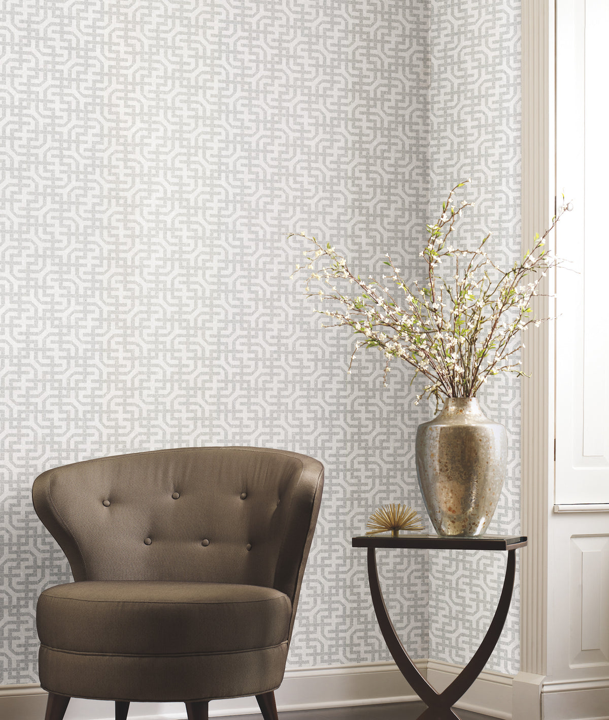 A brown, tufted chair sits beside a small table with a silver vase holding white blossoming branches. The wall is adorned with York Wallcoverings’ Ronald Redding Dynastic Lattice Wallpaper, featuring a stylish gray geometric pattern, while light streams in from a window on the right.