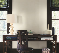 A wooden desk with books, a round lamp, a geometric vase, a black bottle, and a chair sits in front of two windows with black shades and a wall finished in Woven Linen Wallpaper by York Wallcoverings.