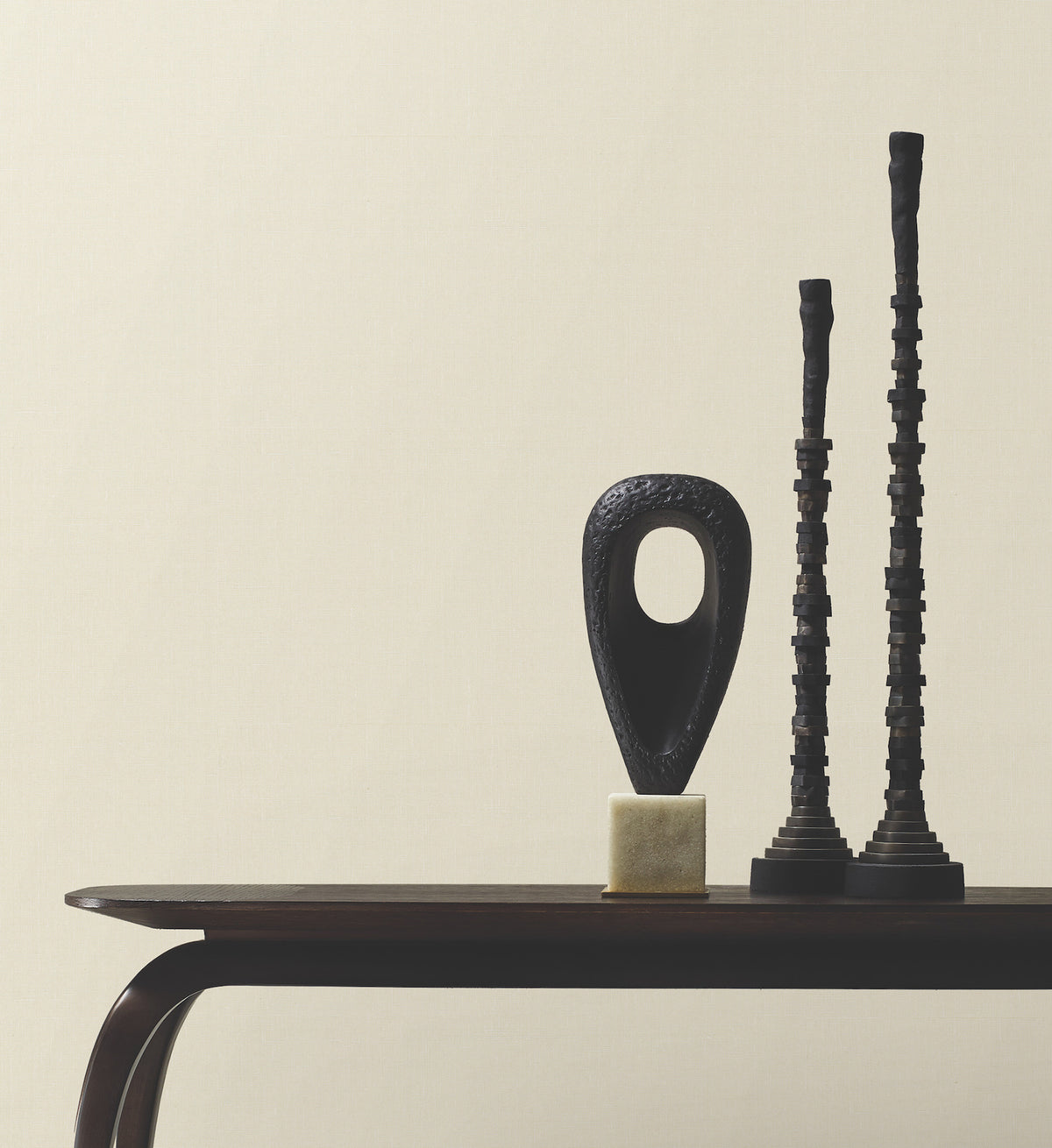 Minimalist photo of a dark wooden table set against a plain wall covered in York Wallcoverings’ Woven Linen Wallpaper, displaying three abstract black sculptures—one oval with a hole, and two tall, narrow, textured columns.