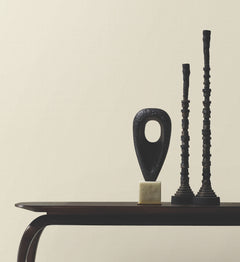 Minimalist photo of a dark wooden table set against a plain wall covered in York Wallcoverings’ Woven Linen Wallpaper, displaying three abstract black sculptures—one oval with a hole, and two tall, narrow, textured columns.