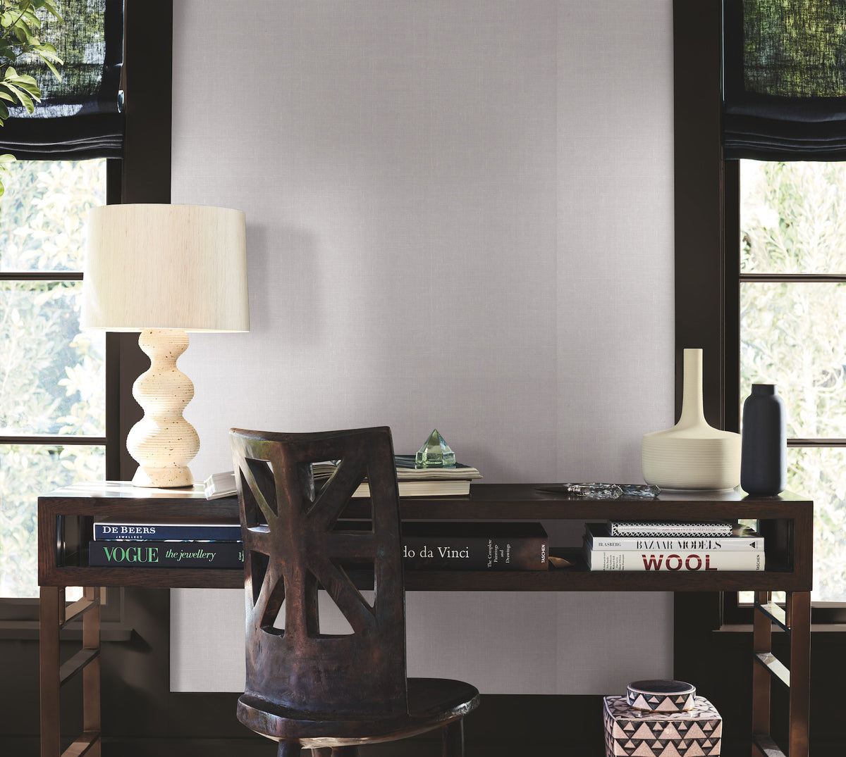 A minimalist wooden desk with art books, a textured lamp, vases, and a geometric chair sits in front of large windows with black shades that let in natural light; the wall is adorned with York Wallcoverings’ Woven Linen Wallpaper for a refined backdrop.