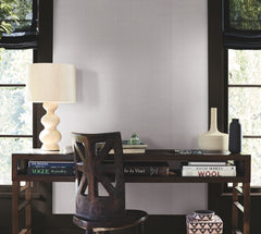A minimalist wooden desk with art books, a textured lamp, vases, and a geometric chair sits in front of large windows with black shades that let in natural light; the wall is adorned with York Wallcoverings’ Woven Linen Wallpaper for a refined backdrop.