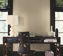 A wooden desk with books, a sculptural lamp, vases, and a decorative box sits in front of a wall covered in York Wallcoverings’ Woven Linen Wallpaper. A chair is pushed under the desk. Two windows with black shades frame the softly textured, neutral wall behind the workspace.