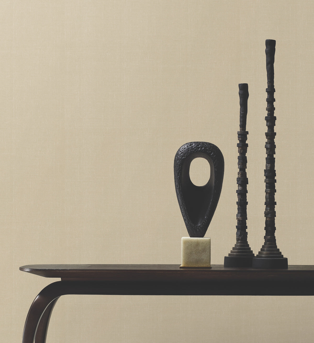 Minimalist tabletop featuring three abstract black sculptures—a hollow oval on a gold base and two tall, stacked cylindrical forms—set against a backdrop of York Wallcoverings’ Woven Linen Wallpaper in a neutral beige tone.