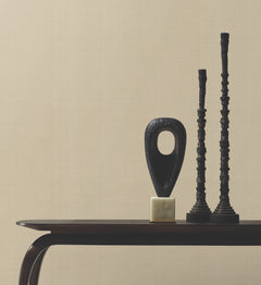 Minimalist tabletop featuring three abstract black sculptures—a hollow oval on a gold base and two tall, stacked cylindrical forms—set against a backdrop of York Wallcoverings’ Woven Linen Wallpaper in a neutral beige tone.