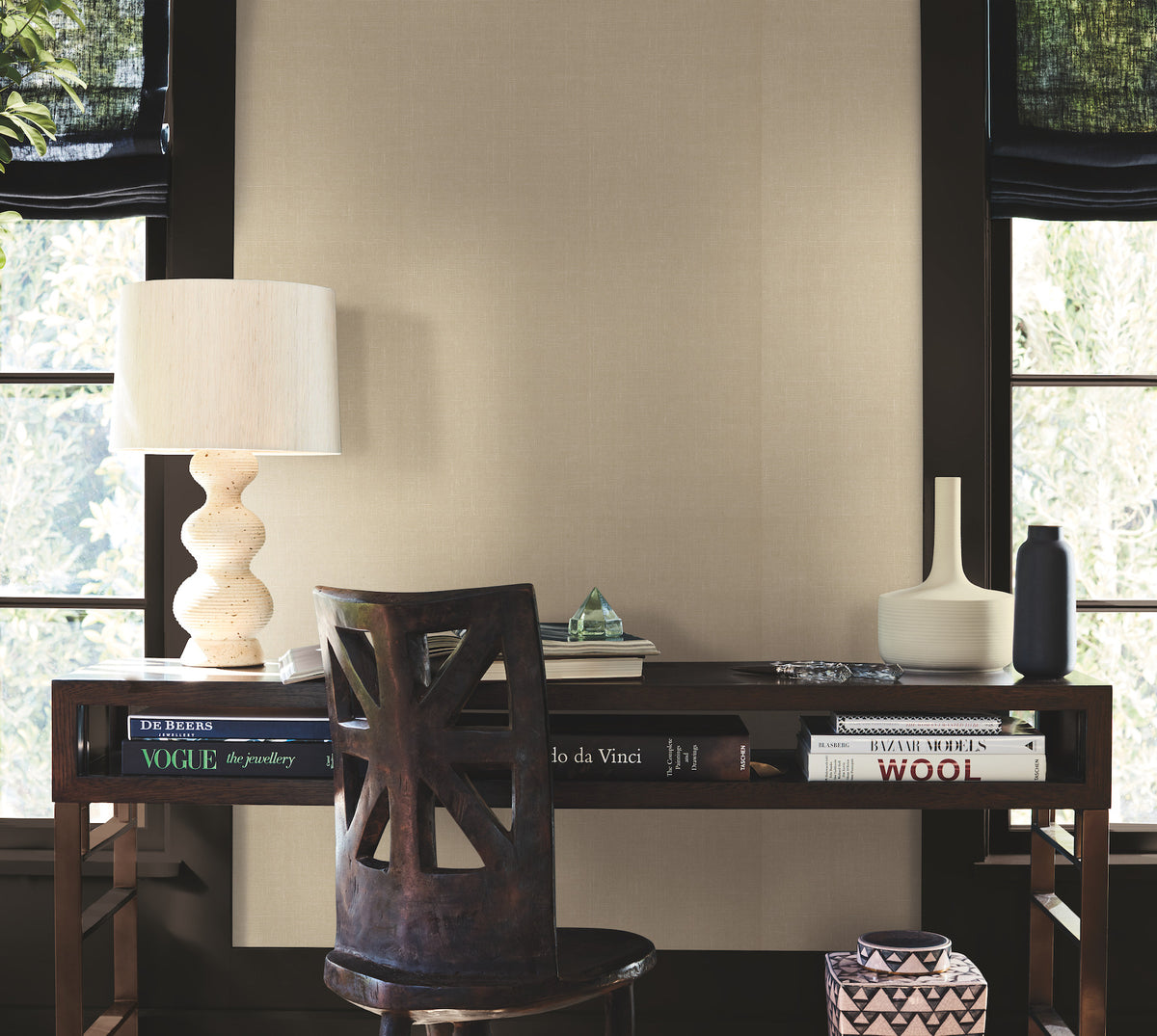 A wooden desk adorned with books, a sculptural lamp, vases, and a geometric decor piece sits in a room featuring Woven Linen Wallpaper by York Wallcoverings. A chair is placed in front of the desk, while two windows with black shades on either side fill the space with natural light.