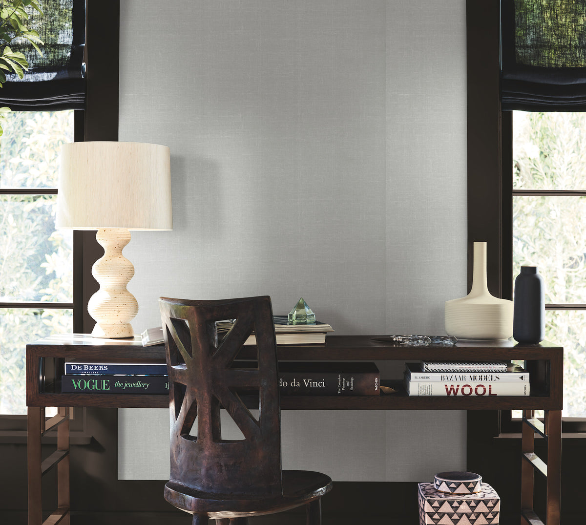 A desk adorned with books, a sculpted lamp, a vase, and a bottle sits before two windows, set against the subtle texture of York Wallcoverings’ Woven Linen Wallpaper. A wooden chair faces the desk as light filters through the trees outside.