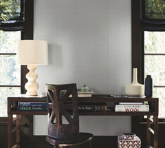 A wooden desk with books, a sculpted lamp, vases, and a chair sits in front of two windows with black roman shades, letting light into the modern minimalist room adorned with Woven Linen Wallpaper by York Wallcoverings.