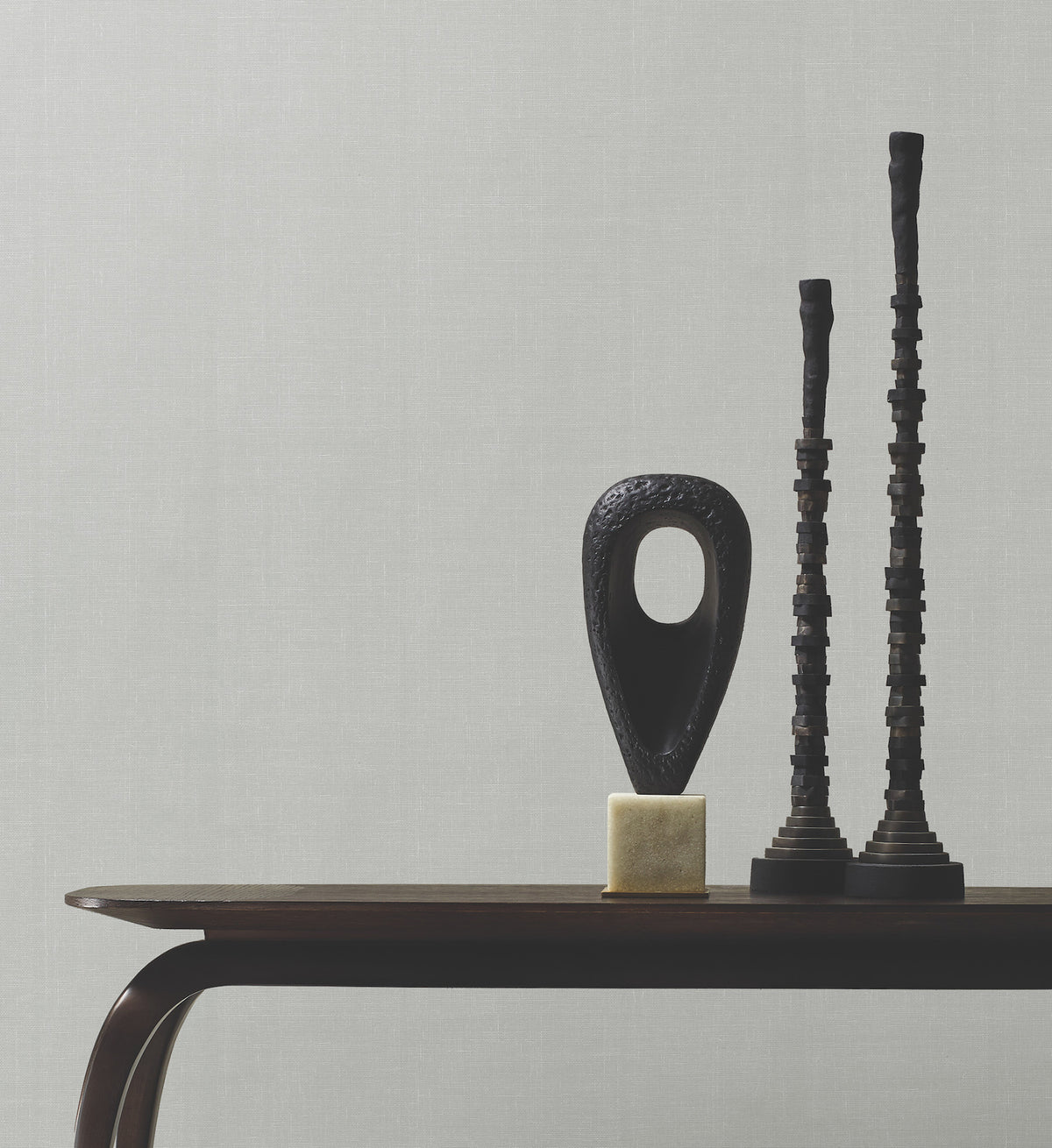 A dark wooden table holds three modern black sculptures—one with a large oval hole on a gold base and two that are tall and textured—set against a plain gray Woven Linen Wallpaper by York Wallcoverings.
