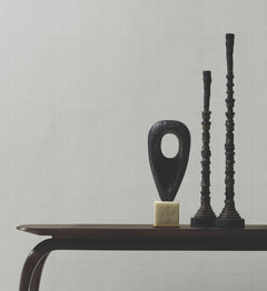 A dark wooden table holds three modern black sculptures—one with a large oval hole on a gold base and two that are tall and textured—set against a plain gray Woven Linen Wallpaper by York Wallcoverings.