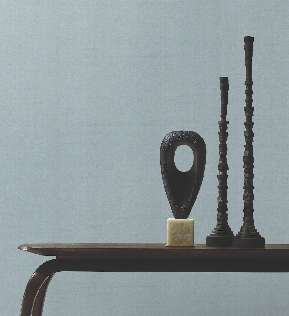 A dark wooden table with abstract black sculptures, including a hollow oval on a cube base and two tall, textured towers, is set against a plain light grey Woven Linen Wallpaper by York Wallcoverings.