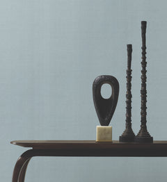 A dark wooden table with abstract black sculptures, including a hollow oval on a cube base and two tall, textured towers, is set against a plain light grey Woven Linen Wallpaper by York Wallcoverings.