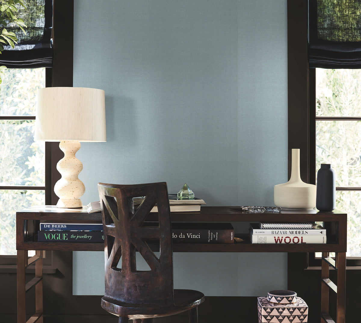 A wooden desk with books, a sculpted lamp, a cream vase, and a dark bottle is positioned in front of two windows. A wooden chair accompanies the desk, and trees can be seen outside. The room features Woven Linen Wallpaper by York Wallcoverings.