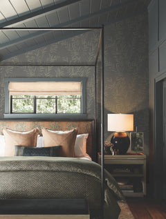 Cozy bedroom with a canopy bed, patterned bedding, pillows, a window with a woven shade, and a nightstand holding a lamp and decor. Walls are covered in Allerton Bamboo Wallpaper in gray with subtle leaf patterns. Light streams in from the window.