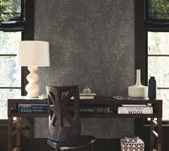 A dark wooden desk with books, a sculpted lamp, vases, and a chair sits in front of windows with black shades. The backdrop features Allerton Bamboo Wallpaper, while sunlight gently filters through the windows.