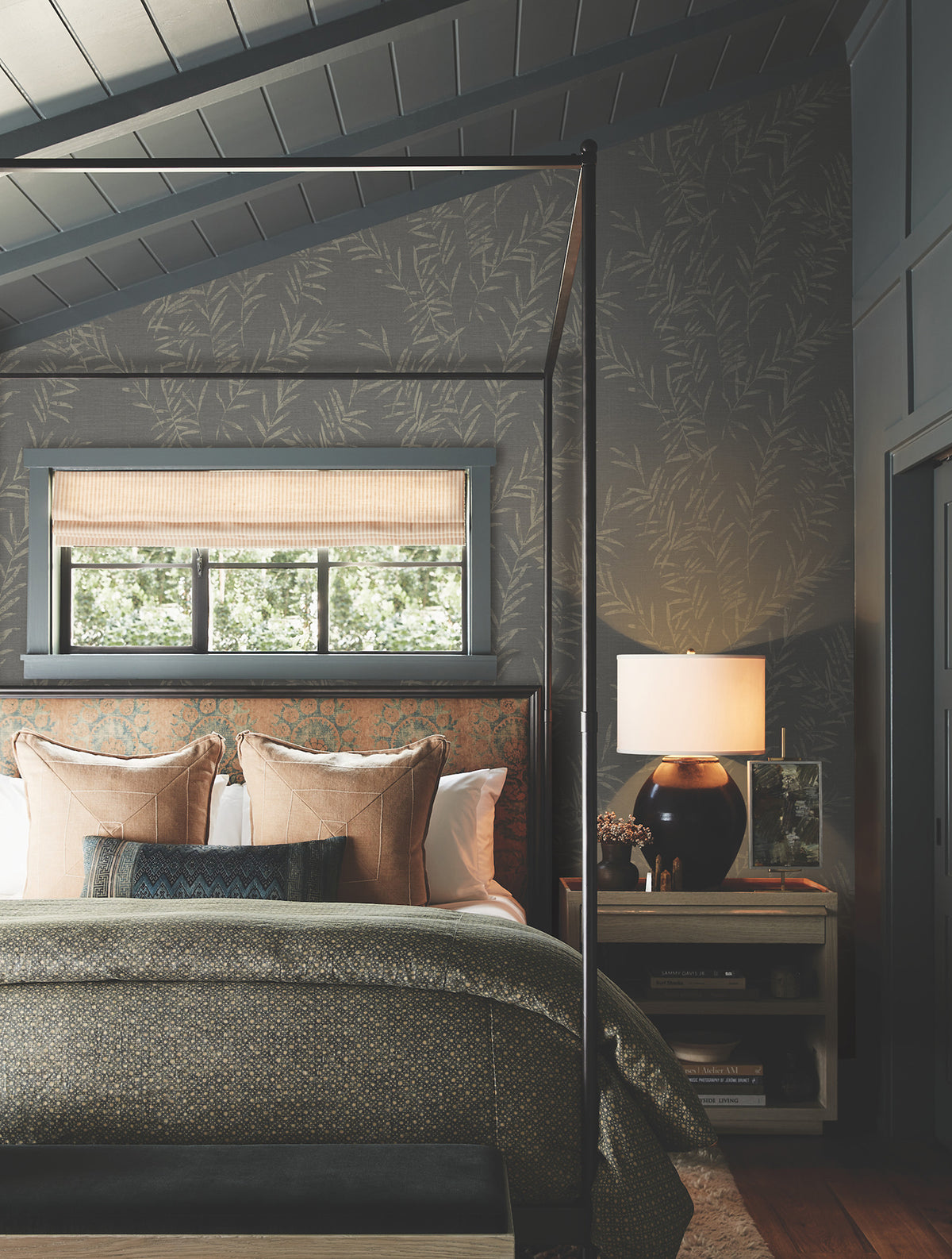 A cozy bedroom featuring a canopy bed with patterned bedding and pillows, a bedside table with a lamp and books, and greenery visible through a window dressed with a bamboo shade. The walls are adorned with Allerton Bamboo Wallpaper, adding a subtle leaf pattern to the space.