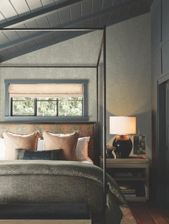 Cozy bedroom with a four-poster bed, earth-toned pillows and blankets, a nightstand with a lamp and books, and a window with light filtering through woven blinds. The accent wall features soft leaf patterns created by Allerton Bamboo Wallpaper.