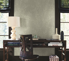 A wooden desk with books, a unique wooden chair, a sculptural lamp, and decorative vases sits in front of two windows with black shades and Allerton Bamboo Wallpaper in the background.