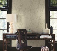 A wooden desk with books, a lamp, and decorative vases sits in front of two windows, backed by Allerton Bamboo Wallpaper, with a dark wooden chair positioned in front.