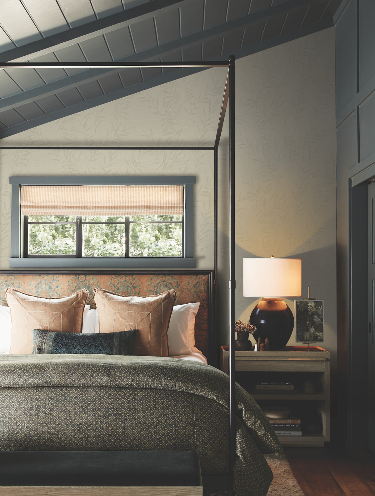 A cozy bedroom featuring Allerton Bamboo Wallpaper, a canopy bed with patterned pillows, a green blanket, a bench at the foot, a window with a woven shade, a nightstand topped with a lamp and a vase of flowers. Light filters in from outside.