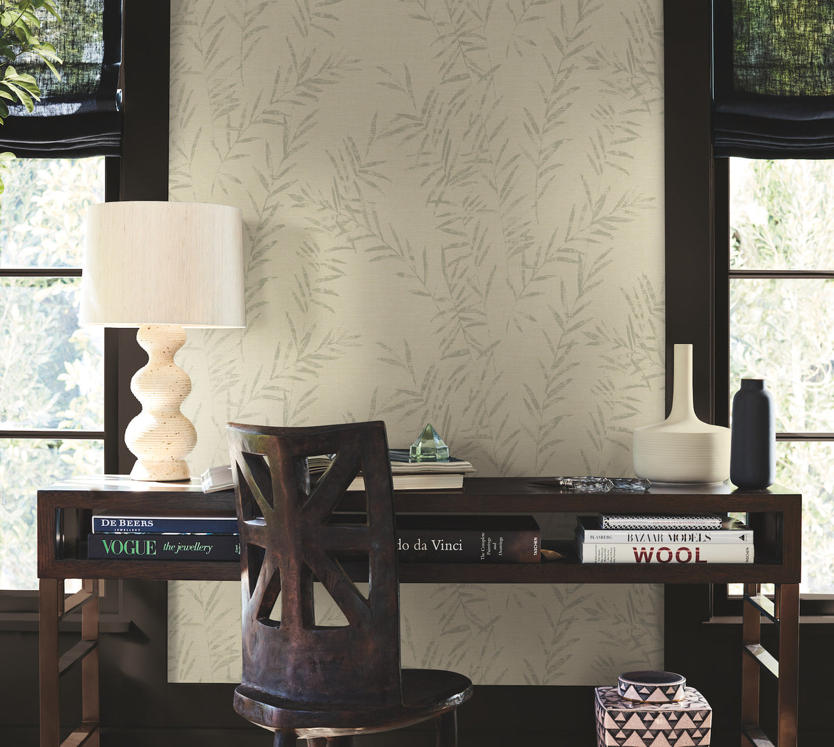 A wooden desk with art books, a cream lamp, vases, and a chair sits in front of windows with black shades and leafy Allerton Bamboo Wallpaper in a cozy, well-lit room.