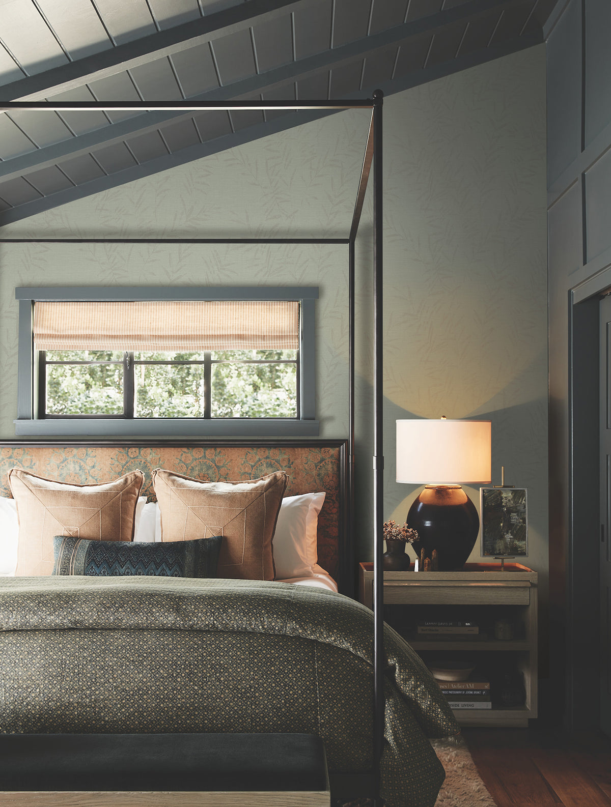 Cozy bedroom featuring a four-poster bed with neutral bedding and decorative pillows, a window dressed in a beige shade, and a nightstand topped with a lamp, books, and a vase—all set beneath a slanted ceiling accented by Allerton Bamboo Wallpaper.