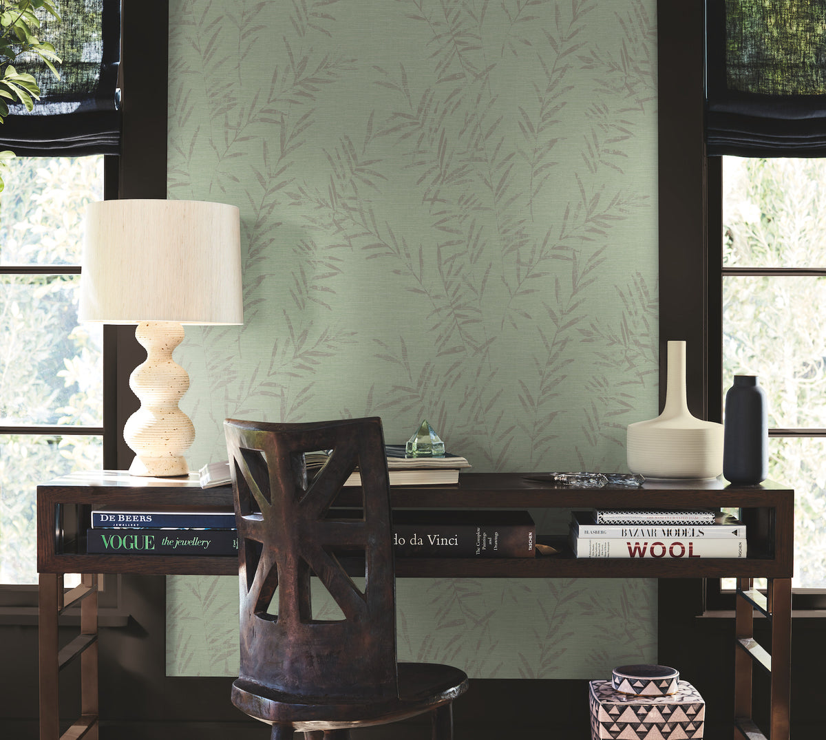 A wooden desk with books, a lamp, and decorative vases sits in front of two windows. A wooden chair is positioned at the desk, and the wall behind is covered in Allerton Bamboo Wallpaper with a light green, leafy pattern.