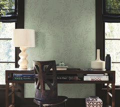 A wooden desk with books, a lamp, and decorative vases sits in front of two windows. A wooden chair is positioned at the desk, and the wall behind is covered in Allerton Bamboo Wallpaper with a light green, leafy pattern.