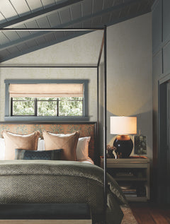 A cozy bedroom featuring Allerton Bamboo Wallpaper, with a canopy bed, patterned pillows, a greenish blanket, a nightstand topped with a lamp and books, and a window adorned with a woven shade welcoming natural light.
