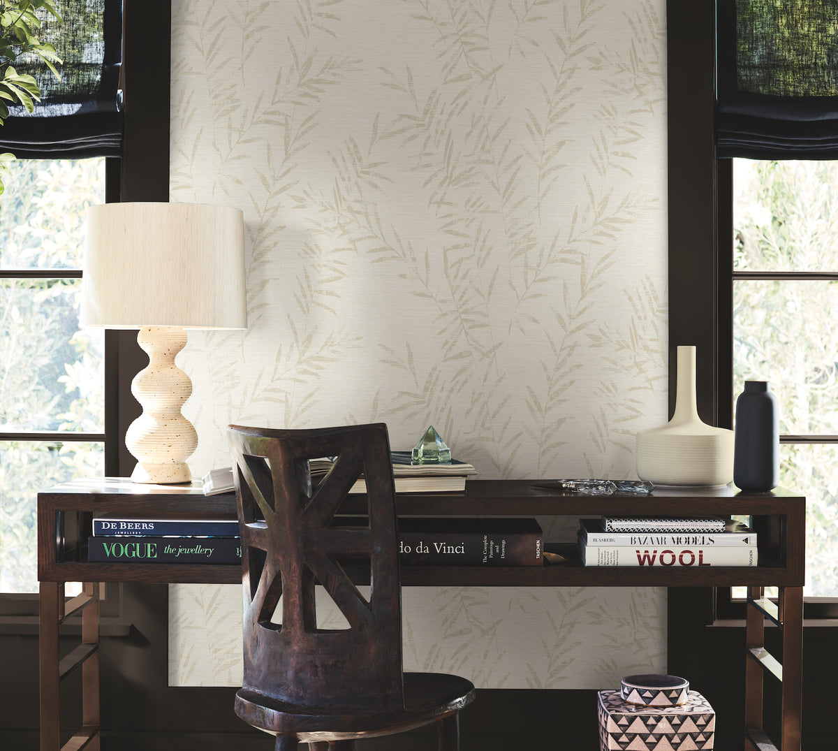 A wooden desk with books, a lamp, and vases sits in front of a wall adorned with Allerton Bamboo Wallpaper. A wooden chair is tucked under the desk, and two large windows provide natural light.