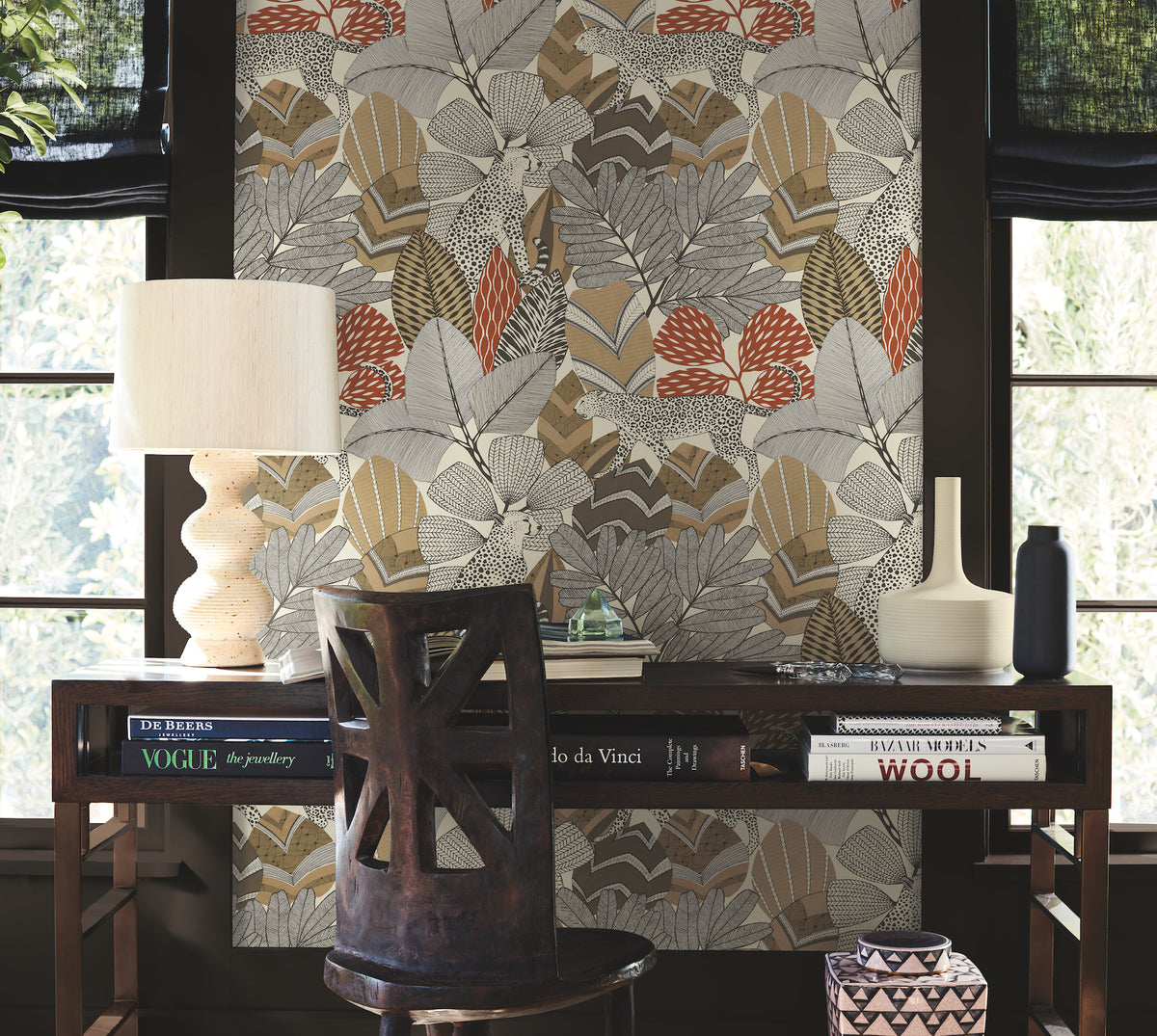 A wooden desk adorned with books, a lamp, and vases sits in front of a wall decorated with Namiri Wallpaper by York Wallcoverings—featuring bold leaf patterns in beige, orange, and gray tones—framed by two windows with black blinds.