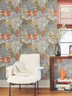 A cozy white armchair draped with a blanket sits beside a small table displaying a vase with branches, books, and a candle. Behind them, the wall is adorned with York Wallcoverings’ Namiri Wallpaper—a bold, floral-patterned design in gray, beige, and orange tones.